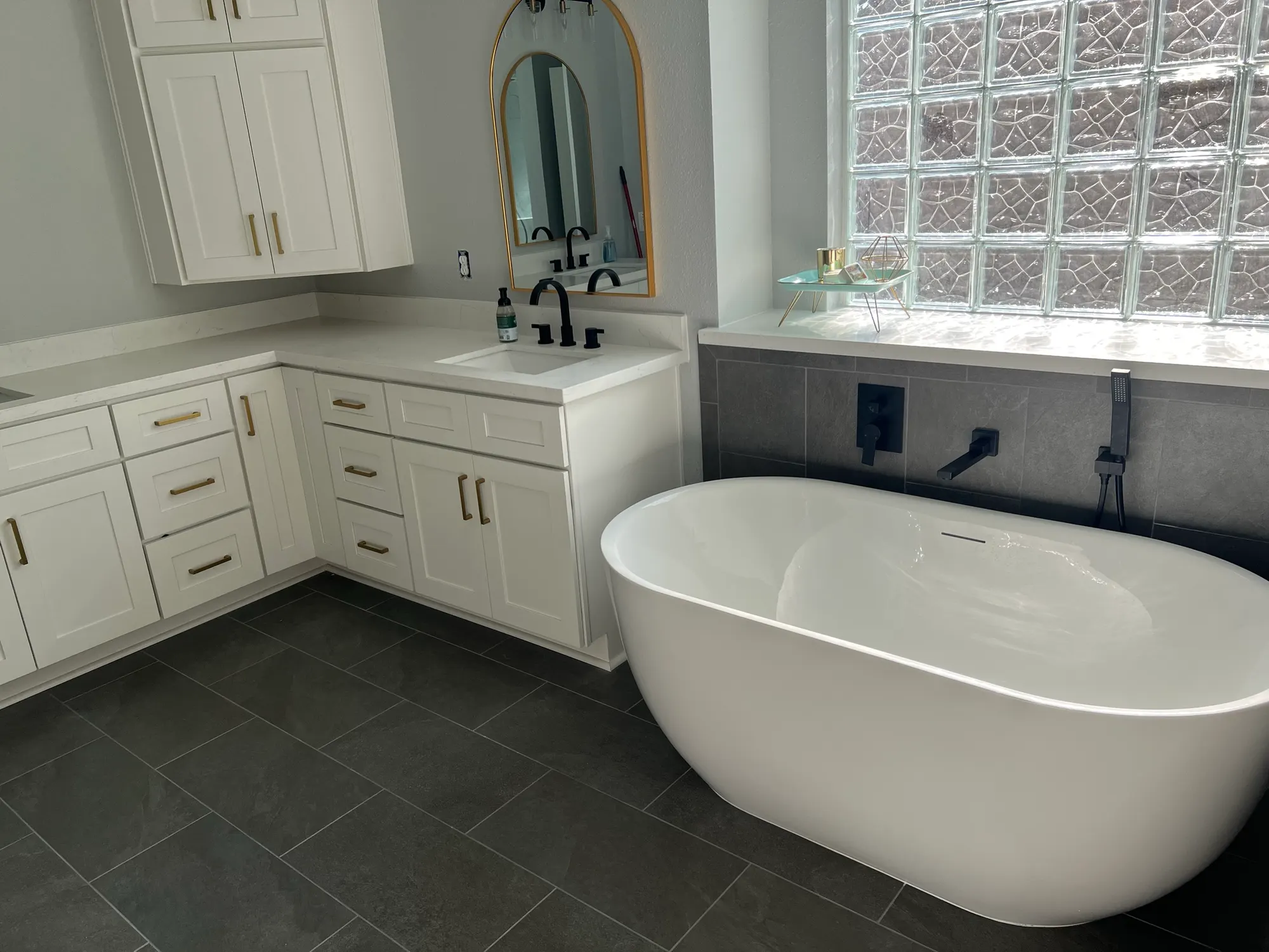 After: freestanding tub under the same glass block window with gray tile floors and a white L-shaped vanity